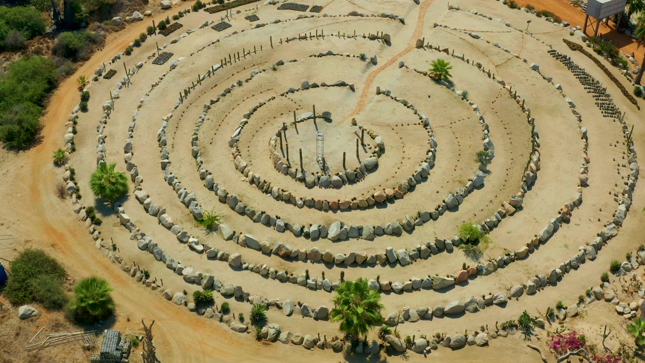 Drone-like top view of concentric stone rows and a sandy path beside shrubs.