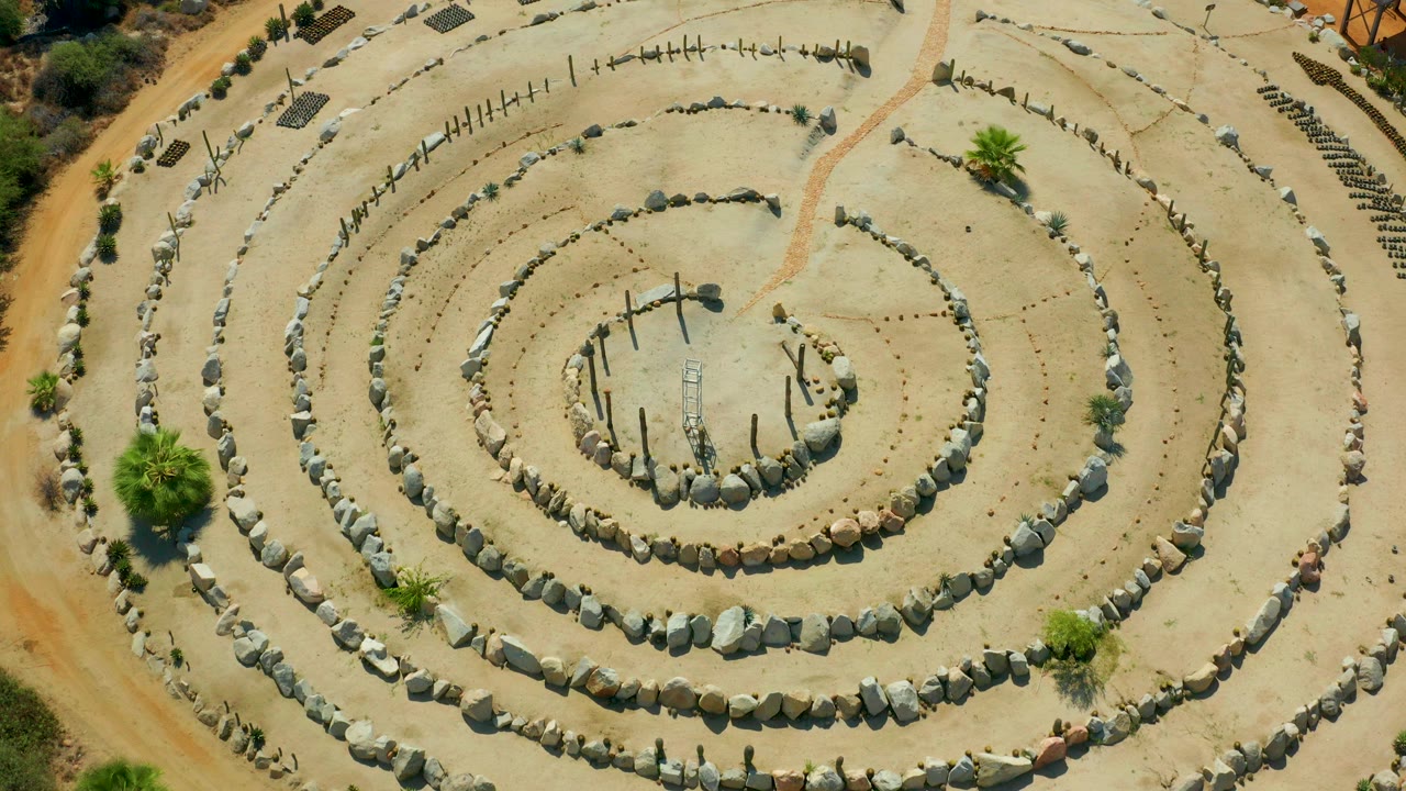 Overhead frame in San Jose Del Cabo, Mexico showing a wide rock spiral in pale sand with sparse desert plants near the edges and no visible people.