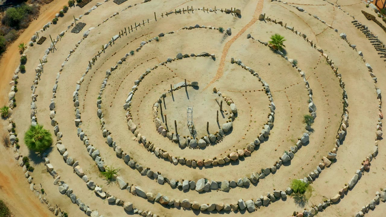 Top-down view in San Jose Del Cabo, Mexico showing the stone spiral, upright sticks in the center, a palm at the lower edge, and no visible people.