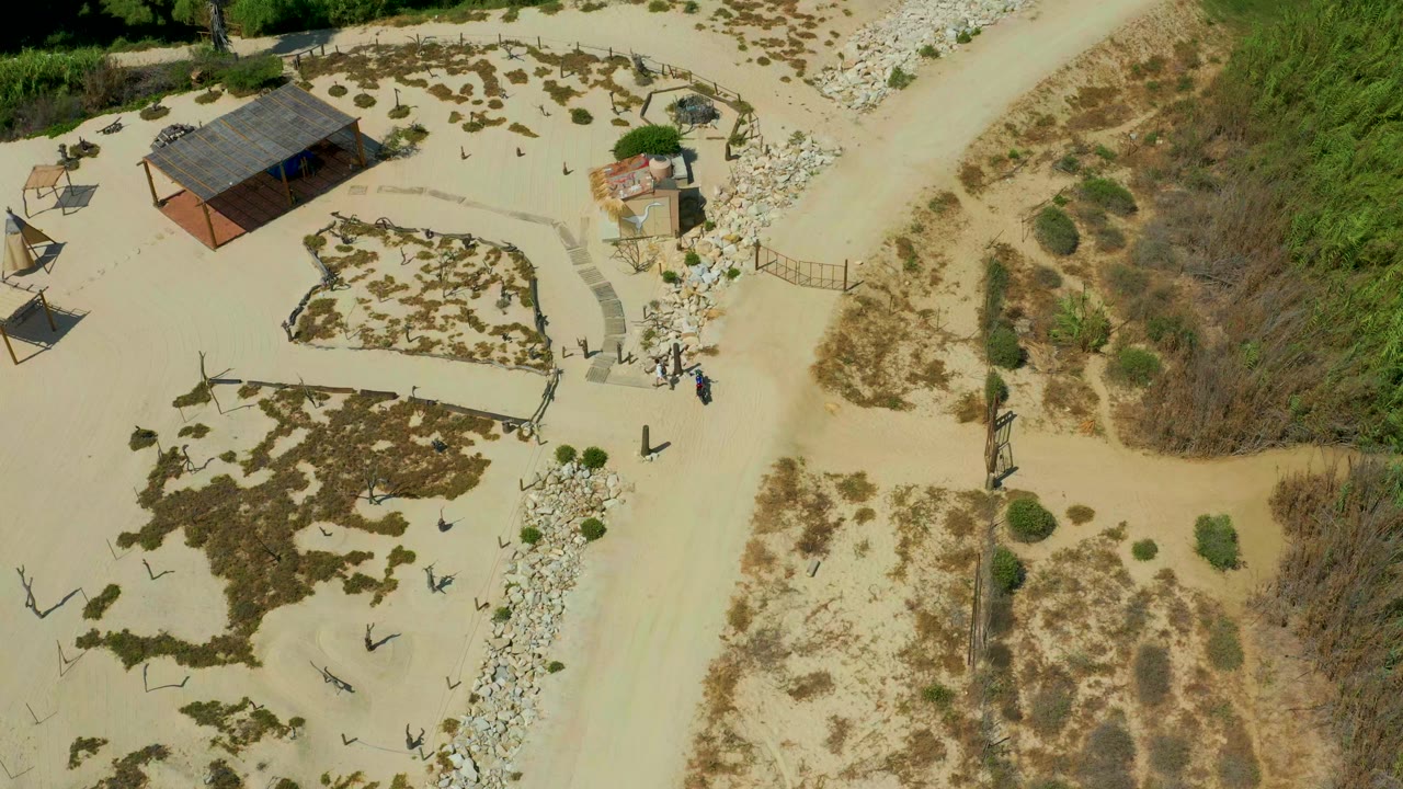High aerial view over a sandy beach area in San Jose Del Cabo, Mexico, showing a small roofed structure, curved paths, rocks, scrub patches, and no visible people.