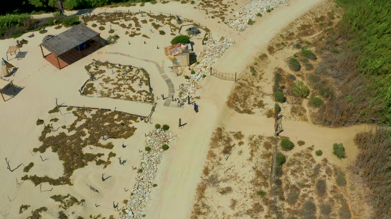 High drone view over San Jose Del Cabo sand and scrub, with a small roofed building, a rectangular sandy enclosure, rock edging, and a pale road beside dry brush, with no visible people.