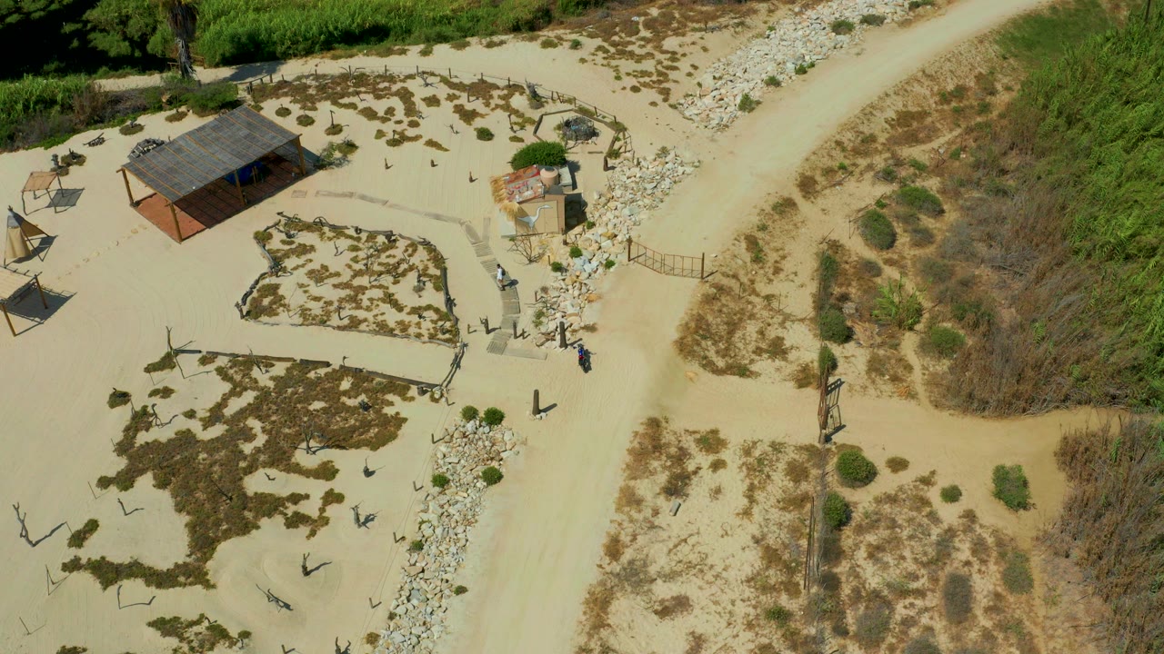 Overhead frame from San Jose del Cabo, Mexico, showing a small shack near pale sandy trails, stone edging, scrub vegetation, and tiny visible people near the path.
