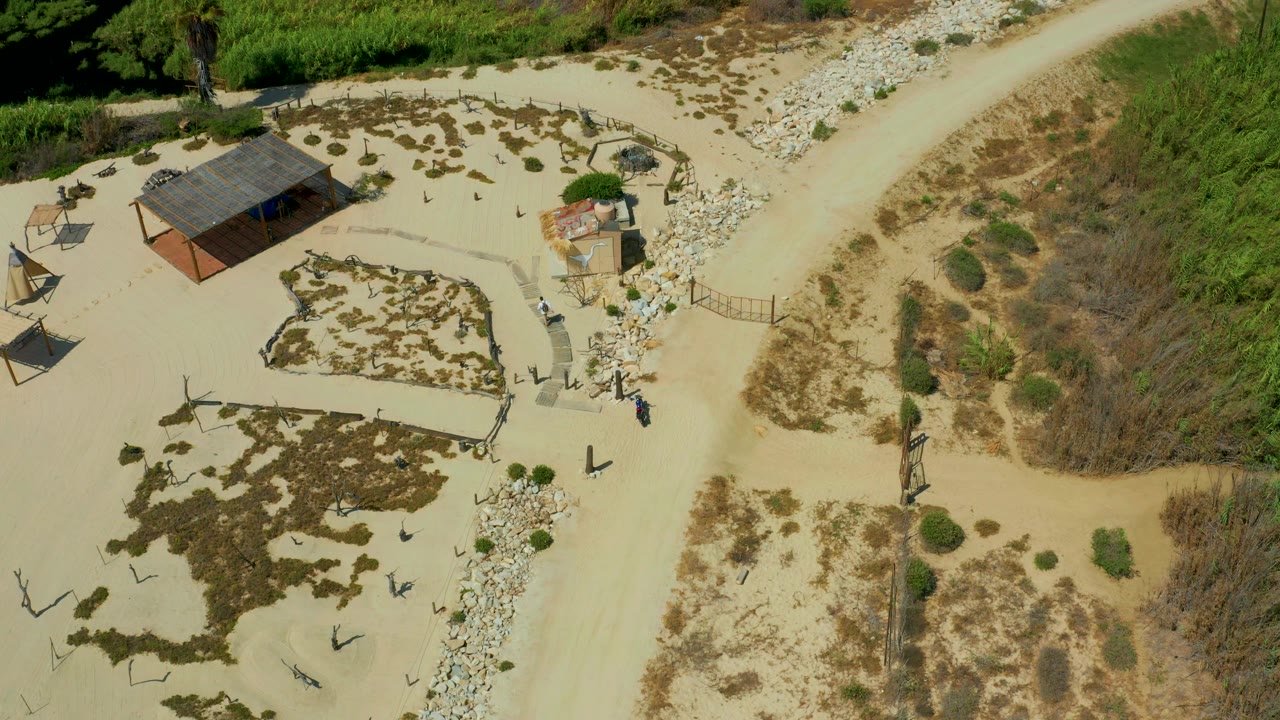 High overhead frame from San Jose del Cabo, Mexico, showing the small shack, a forked sandy path, green brush along the top edge, and tiny figures near the right-side walkway.