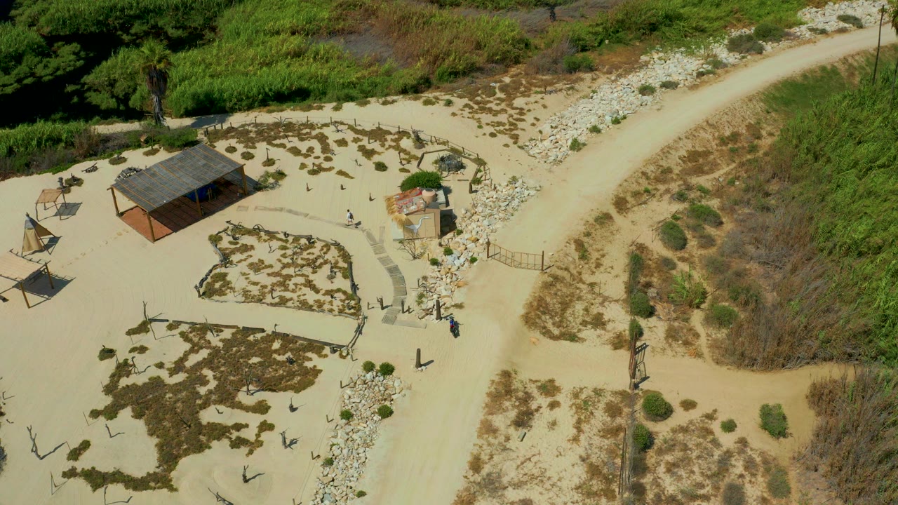Overhead frame in San Jose Del Cabo, Mexico showing pale sand paths, a roofed shelter, shade structures, scrub, rocks, and tiny people near the walking areas.