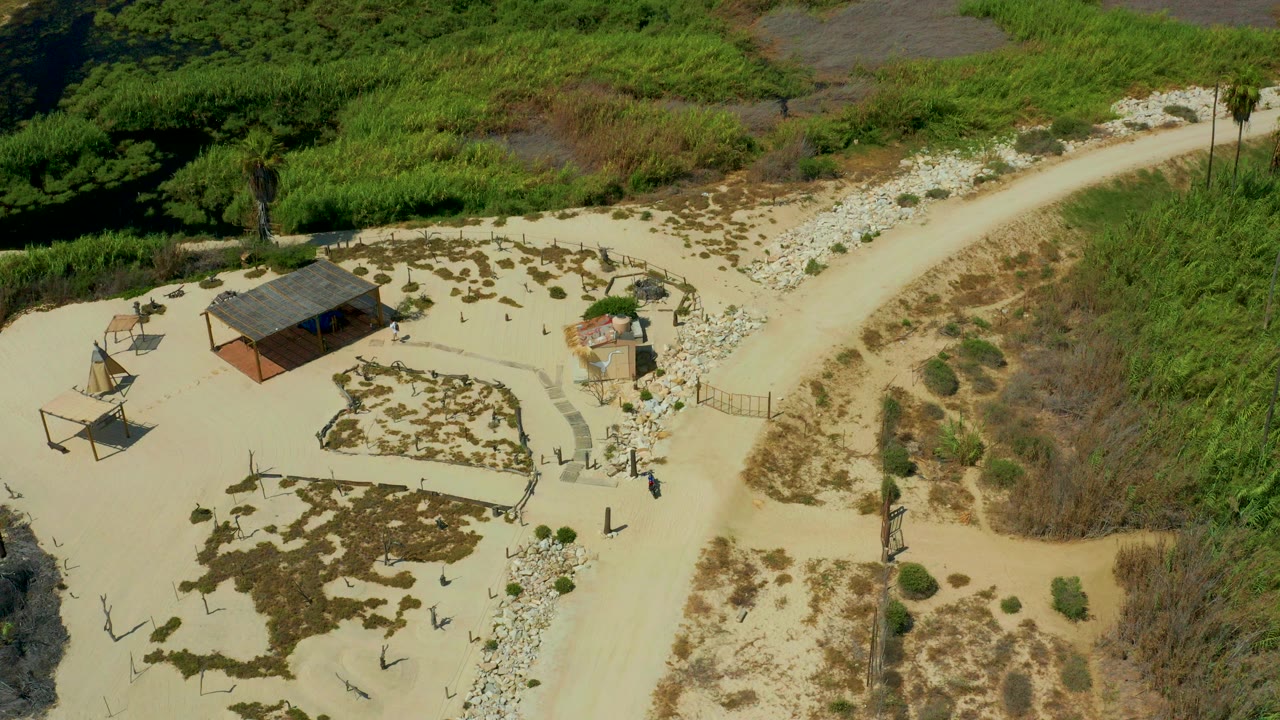 Aerial frame over San Jose Del Cabo, Mexico showing a small roofed structure in a sandy clearing, shade frames, a curved dirt road, green brush, and no visible people.