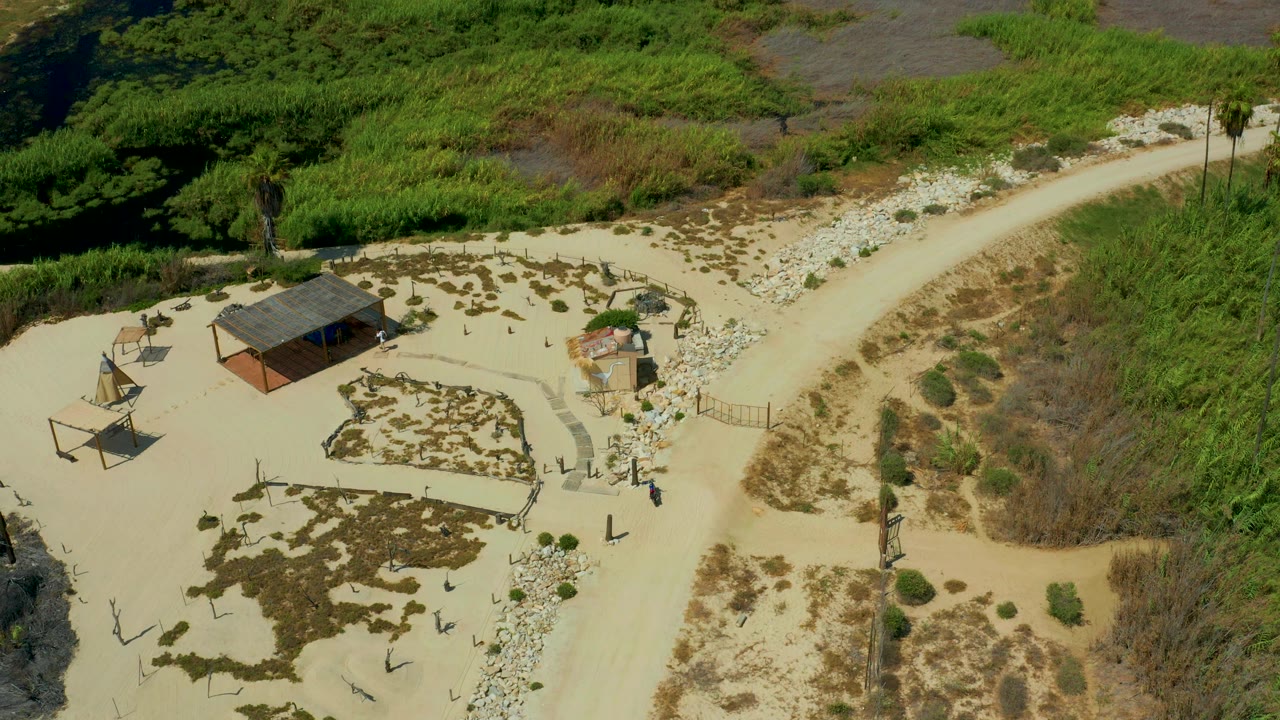 Aerial view over San Jose Del Cabo, Mexico showing a hut-like roofed building, two shade-frame structures, scattered rocks, tan sand, green brush, and no visible people.