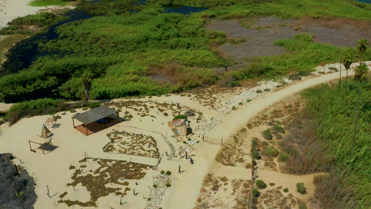High aerial view in San Jose Del Cabo, Mexico, showing a small roofed shack, pale sandy ground, a curving rural trail, green wetland plants, background water, and no visible people.