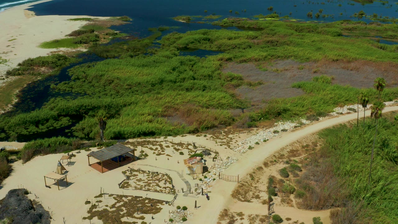 Aerial view in San Jose Del Cabo, Mexico, showing pale sand, green marsh grass, dark lagoon channels, blue water at the top edge, and no visible people.