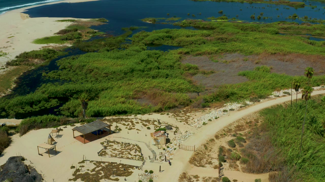 Aerial view in San Jose Del Cabo, Mexico, with a curving sandy track along marsh vegetation, blue water in the background, a small shelter near the lower frame, and no visible people.
