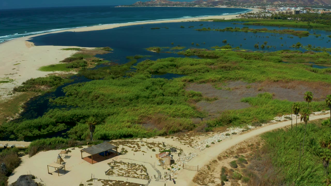 San Jose del Cabo aerial frame with green marshland filling the foreground, a sandbar and dark lagoon in the middle, ocean waves beyond, distant town buildings, and no visible people.