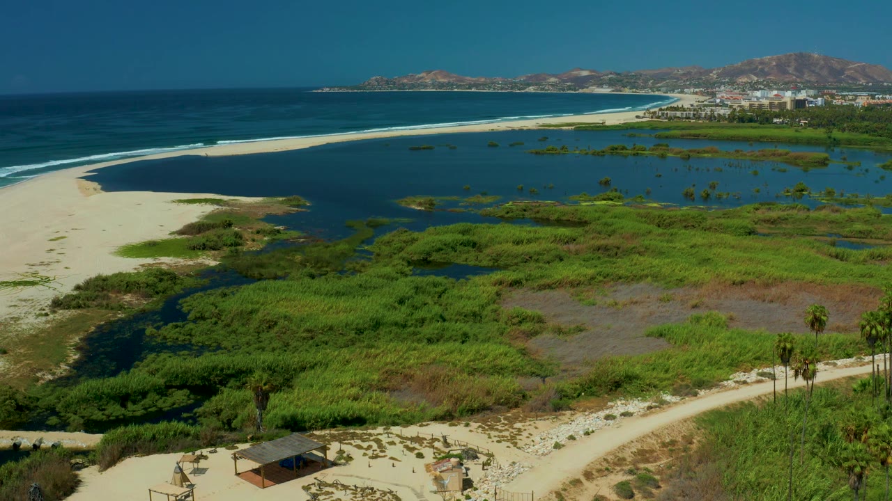 San Jose Del Cabo aerial still showing a broad lagoon, green wetland patches, a long pale beach, open blue sea, distant buildings at the shoreline, hills, and no visible people.