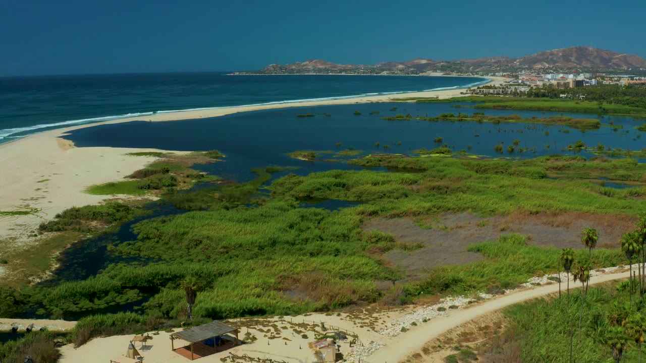 Aerial view of sandy path, palms, lagoon, beach, ocean, and distant hills.