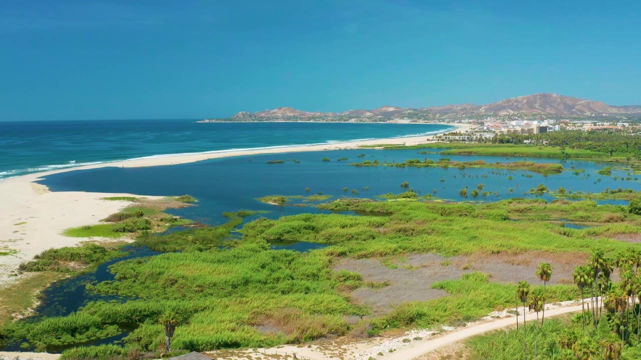 At 0:01, the San Jose del Cabo aerial view keeps the blue sea and white surf line across the top of frame, with sandbar, lagoon, green wetlands, distant buildings, hills, and no visible people.