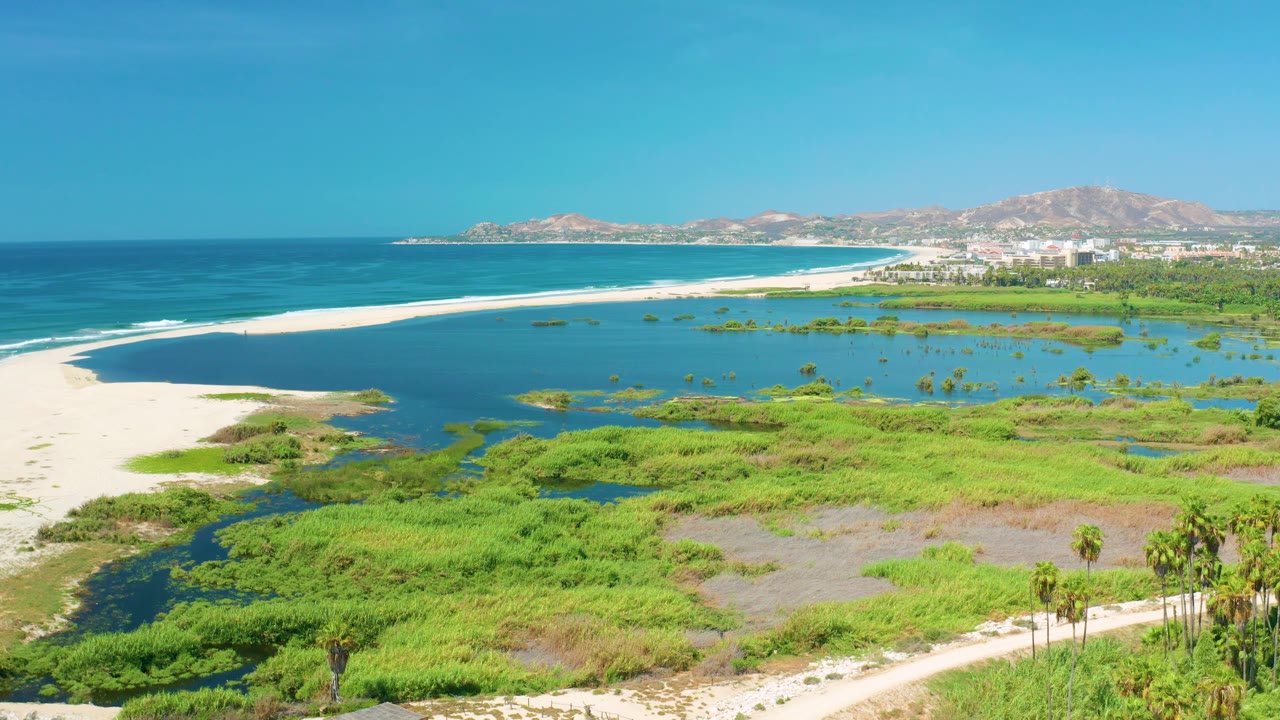 At 0:03, bright light covers the San Jose del Cabo wetlands and lagoon, with the beach and surf curving toward distant buildings and hills, and no visible people.