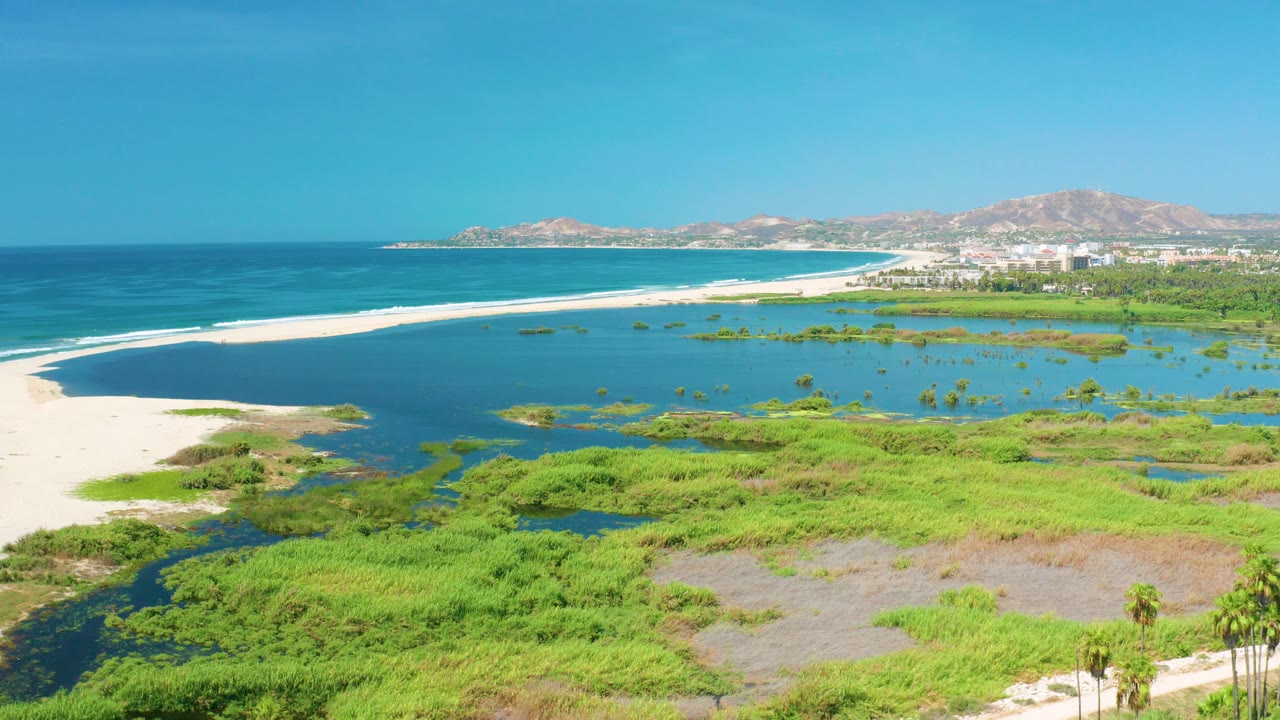 Opening aerial frame over San Jose del Cabo, Mexico, showing a long white sandbar dividing deep ocean from shallow lagoon water, with green wetlands below and no visible people.