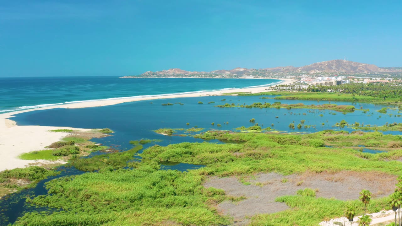 At 0:02, the San Jose del Cabo coastline appears from above with the white sandbar stretching left to right, wetland pools and vegetation below, distant buildings, mountains, and no visible people.