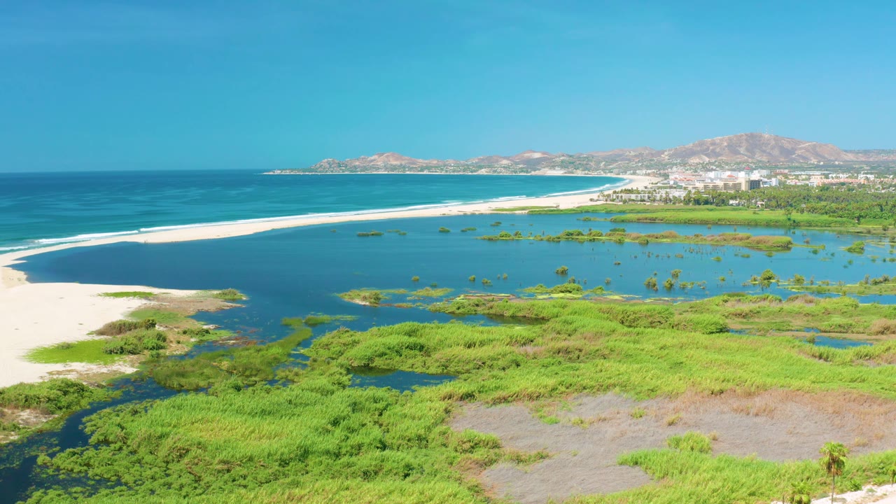 At 0:03, the aerial composition places the white sandbar and ocean surf line across the middle, with green wetland textures in front, San Jose del Cabo buildings beyond, and no visible people.