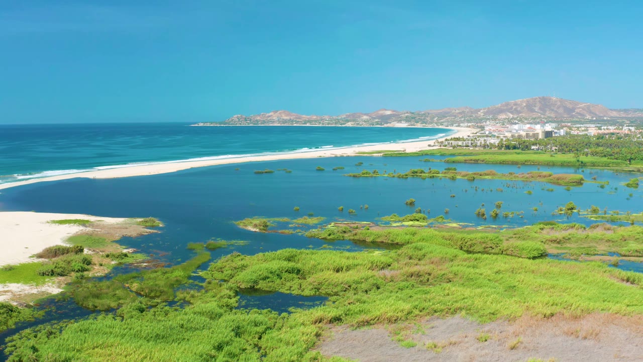 Aerial view in San Jose Del Cabo, Mexico, with a broad lagoon filling the center, white waves along the sandbar, green coastal vegetation below, distant shoreline buildings, and no visible people.