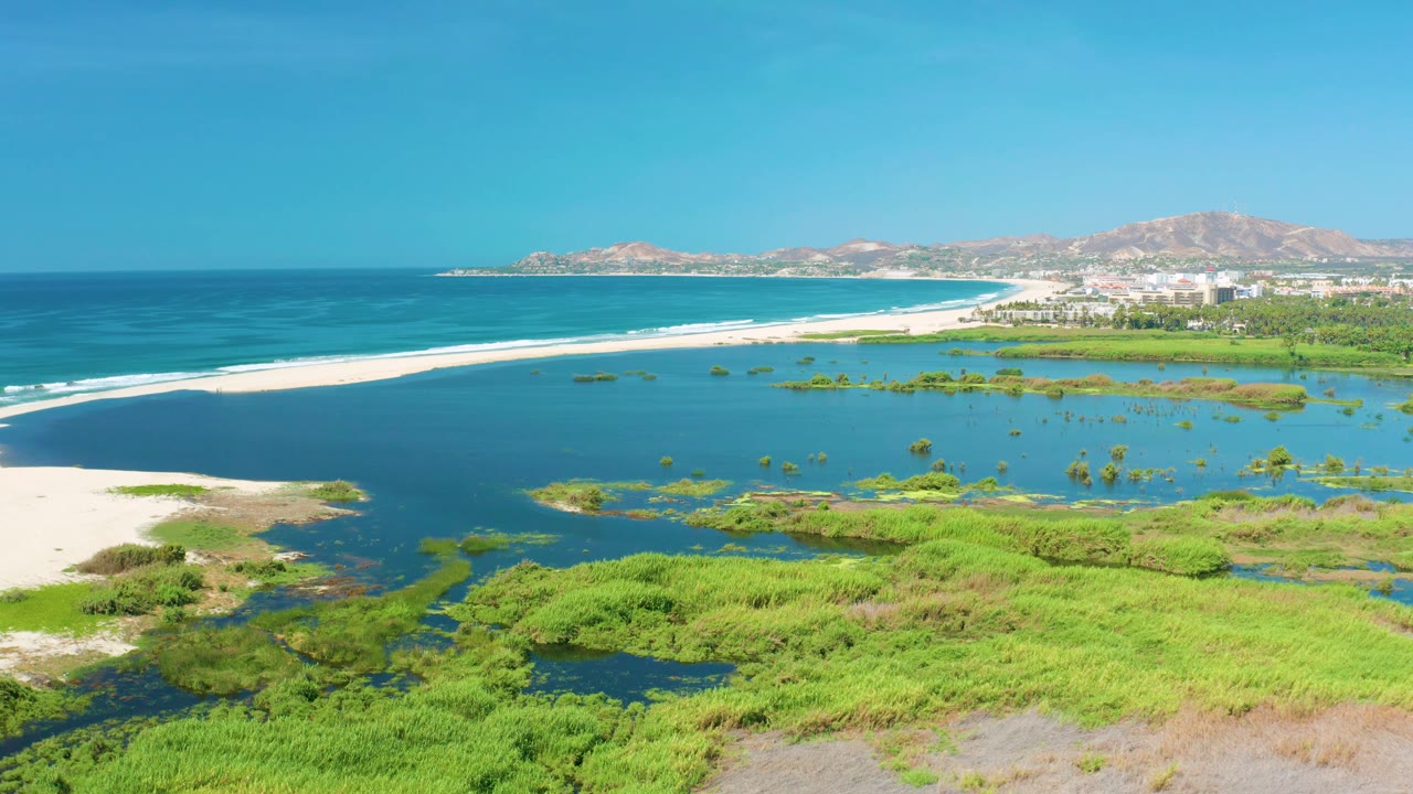 San Jose Del Cabo aerial frame showing the lagoon and marsh spread across the foreground, the white beach separating lagoon from ocean, distant hills, coastal buildings, and no visible people.