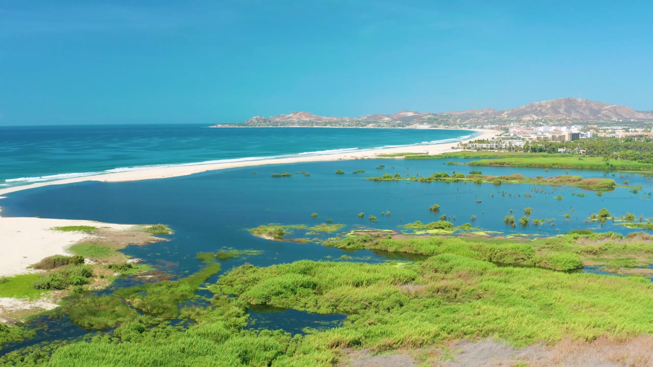 Aerial frame from San Jose Del Cabo, Mexico, showing the shoreline from a slightly later moment, with turquoise lagoon water, white sandbar, open ocean, distant town structures, hills, and no visible people.