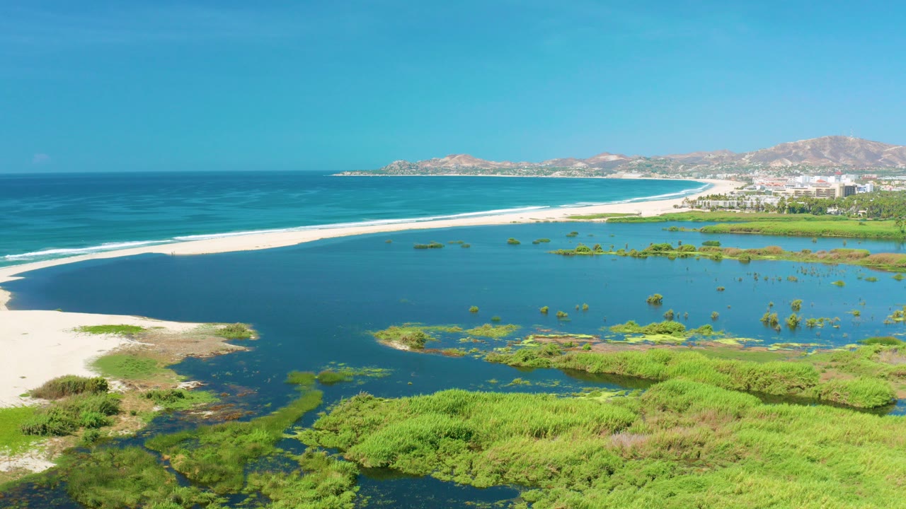 Aerial view in San Jose del Cabo, Mexico, with green marsh in the foreground, a dark lagoon, white beach, blue ocean, distant resort buildings, and no visible people.