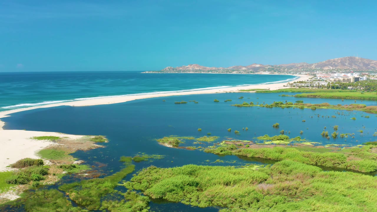 Wide aerial view in San Jose del Cabo, Mexico, with a long white beach dividing the dark lagoon from the blue ocean, backed by low hills and no visible people.