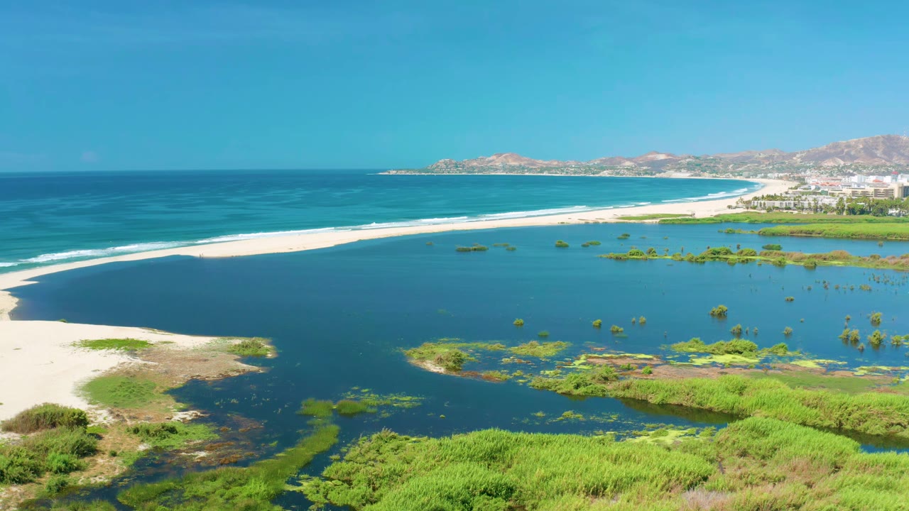 Wide aerial frame of San Jose Del Cabo coastline with the lagoon filling the foreground, low green vegetation at the bottom edge, mild waves along the beach, and no visible people.