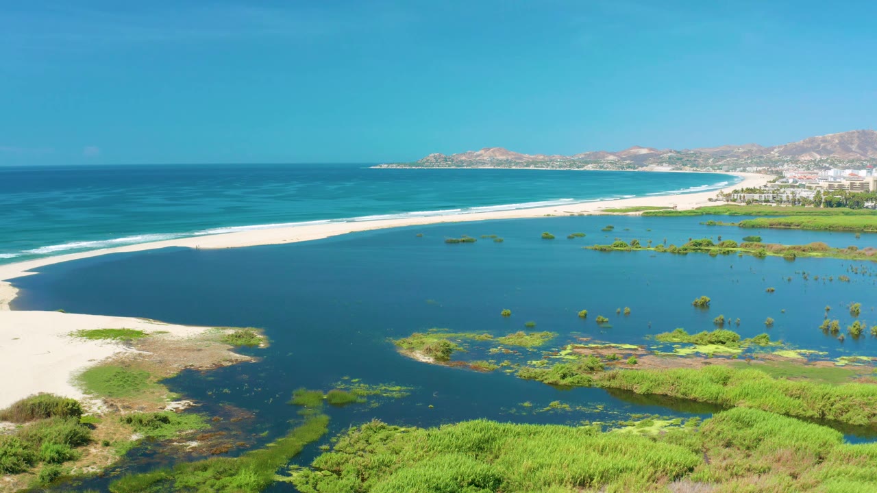 San Jose Del Cabo aerial view with bright turquoise ocean, a long white sand beach, dark lagoon water broken by green plant patches, distant shoreline buildings, and no visible people.