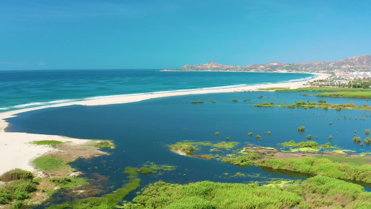 Elevated San Jose Del Cabo coast view with lagoon water across the lower frame, a small sandy point at left, white beach and surf in the middle, hills and buildings in the distance, and no visible people.