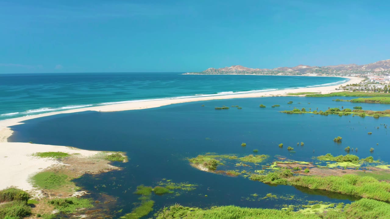 Wide aerial frame from San Jose Del Cabo, Mexico, showing a curved white sand bar between turquoise ocean and darker lagoon water, with wetlands below and no visible people.