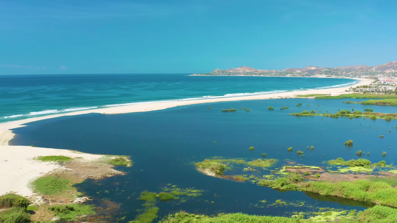 Elevated view in San Jose Del Cabo, Mexico, with the ocean horizon across the top, a pale sand strip through the center, lagoon water and green patches below, and no visible people.
