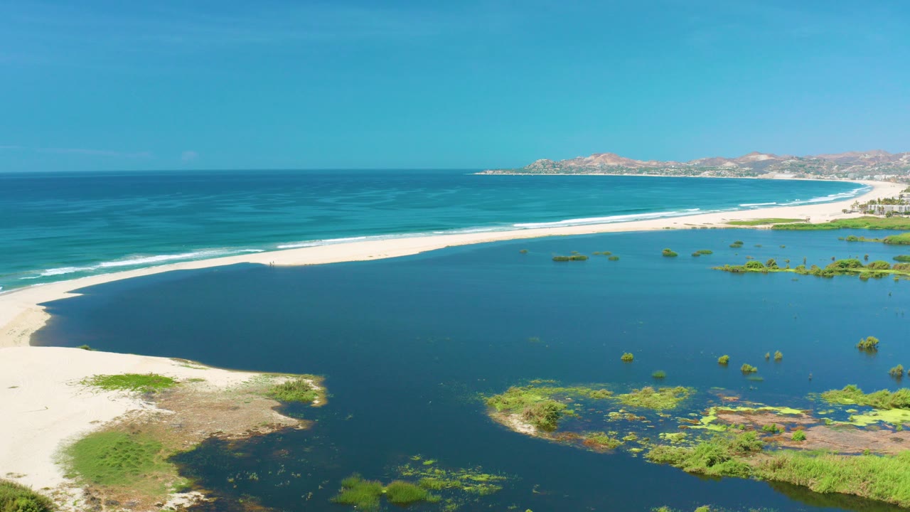 Aerial frame from San Jose Del Cabo showing a pale sandbar between dark lagoon water and turquoise ocean surf, with low hills on the horizon and no visible people.