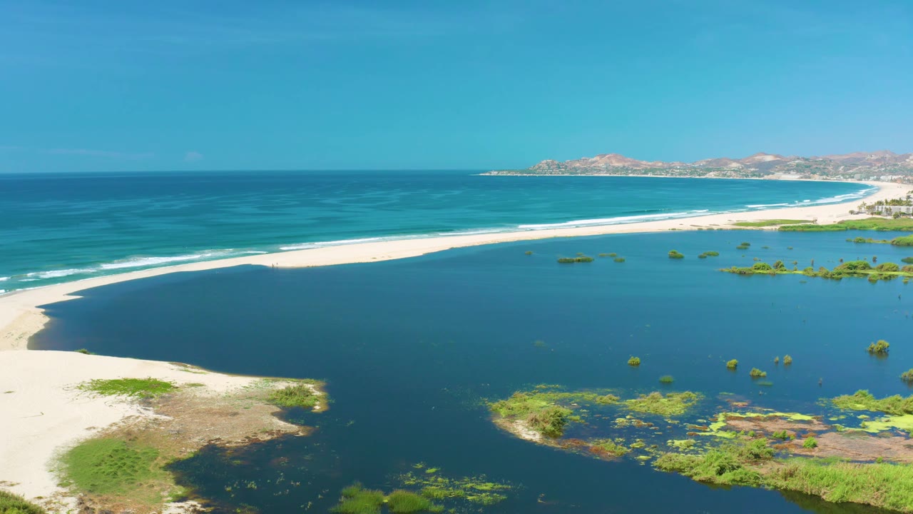 The frame shows San Jose Del Cabo coastline from above, with white sand at left, blue-green lagoon water in front, ocean surf behind, and no visible people.