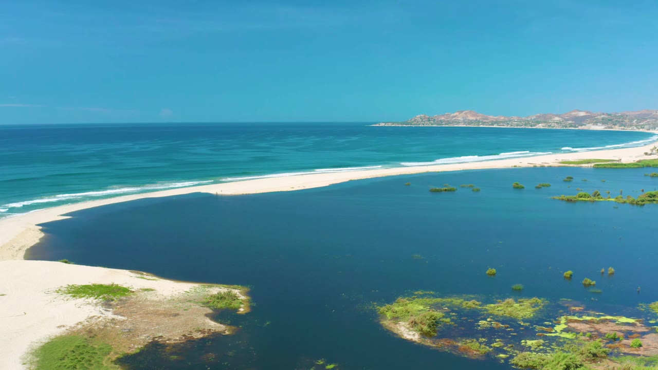Wide drone view of curving beach, blue sea, and shallow green patches near shore.