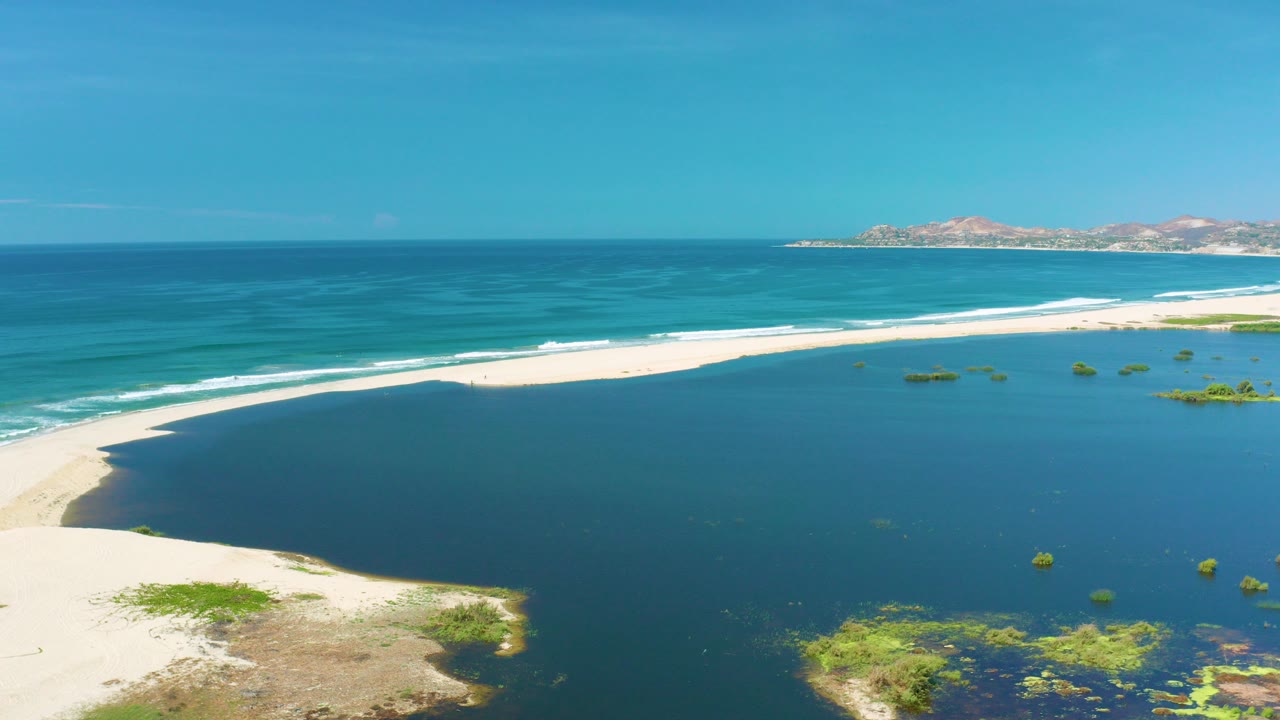 Aerial frame over San Jose Del Cabo with dark blue lagoon water in the foreground, a pale sandbar, turquoise surf beyond it, distant hills on the horizon, and no visible people.