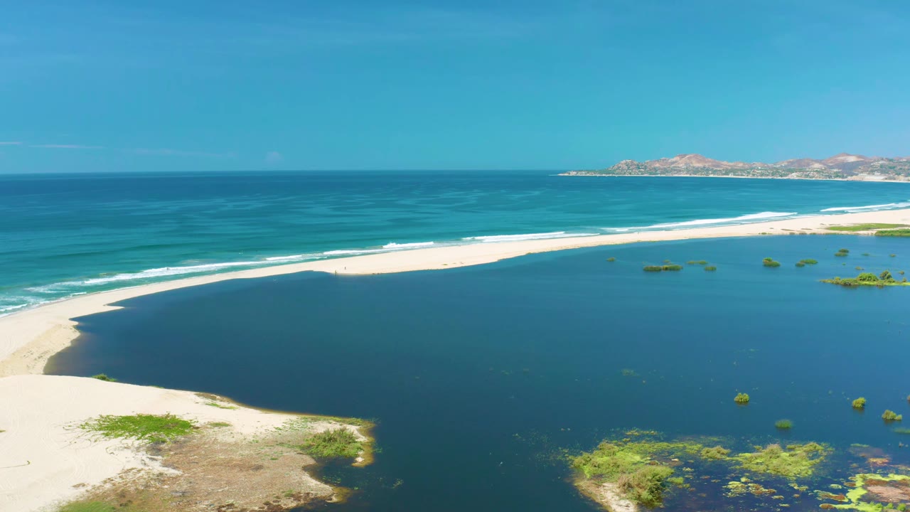 Aerial view in San Jose Del Cabo showing the sandbar curving between darker lagoon water and brighter ocean waves, with low hills in the background and no visible people.