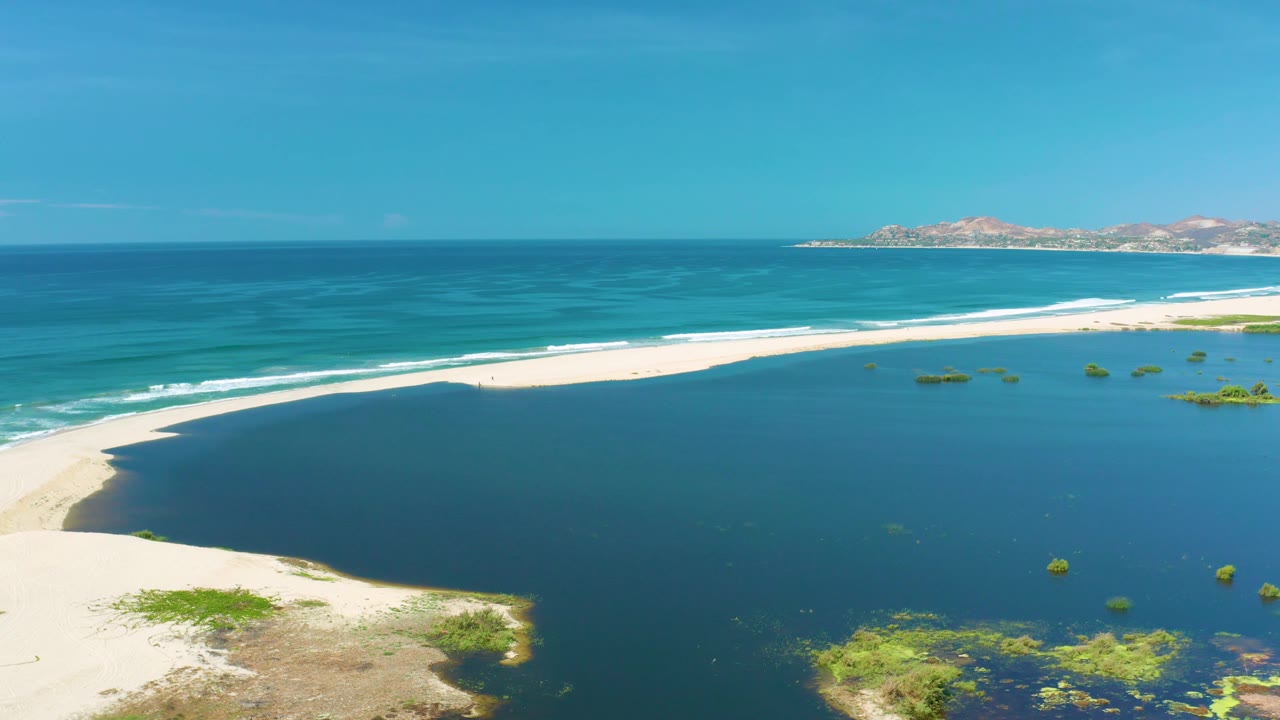 High coastal frame from San Jose Del Cabo with a white sand spit running across the scene, dark water below, rolling surf above, distant dry hills, and no visible people.