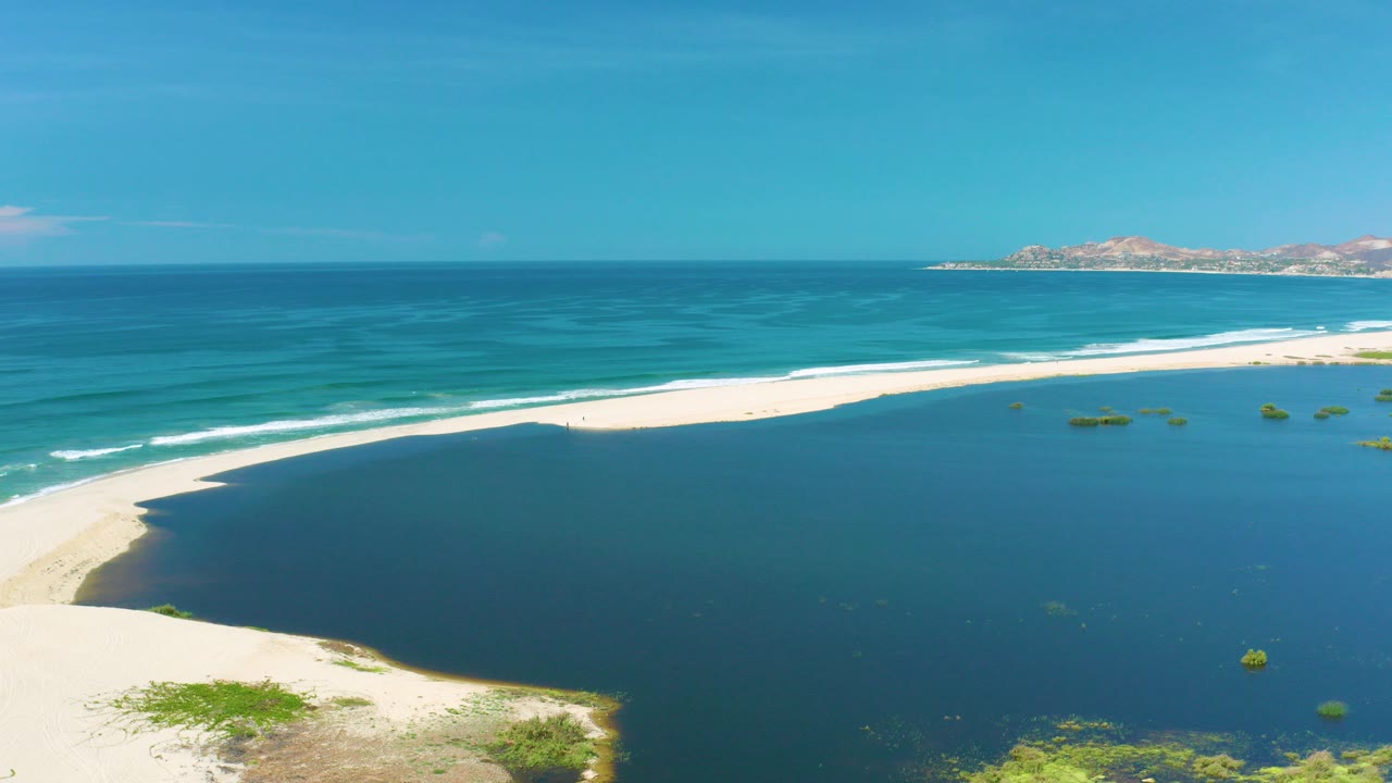 Aerial view in San Jose Del Cabo, Mexico, showing a curved white sand beach, dark blue inlet water, turquoise ocean bands, distant rocky land, and no visible people.