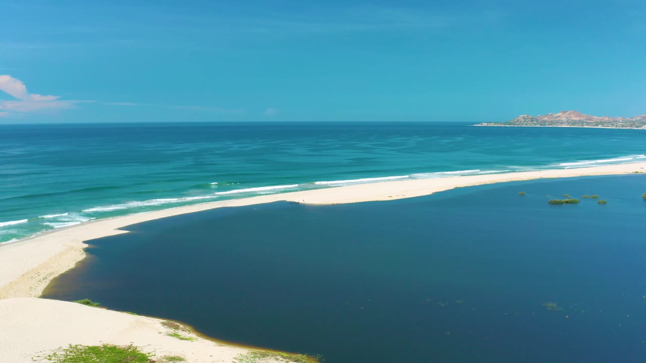 At 0:02, the San Jose Del Cabo shoreline remains wide in frame, with dark lagoon water below, a bright sand curve, ocean bands, a rocky distant coast, and no visible people.