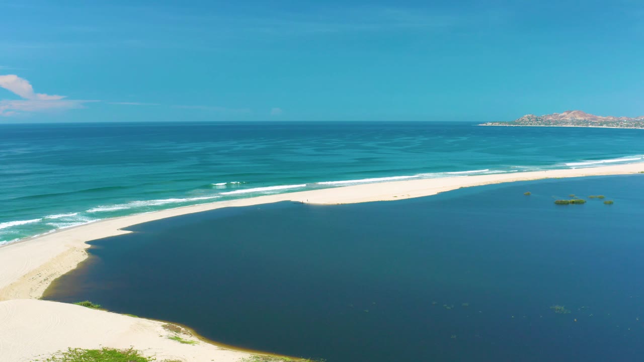 Mid-clip frame over San Jose Del Cabo, Mexico, showing the lagoon as a broad dark shape below the beach, multiple surf bands offshore, distant land to the right, and no visible people.