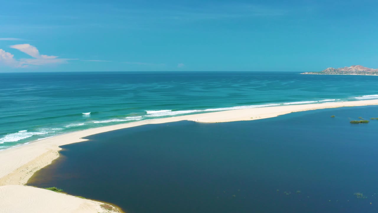 San Jose Del Cabo drone view showing the long sandbar stretched across the frame, gentle white wave lines, open blue ocean, a small rocky horizon feature, and no visible people.