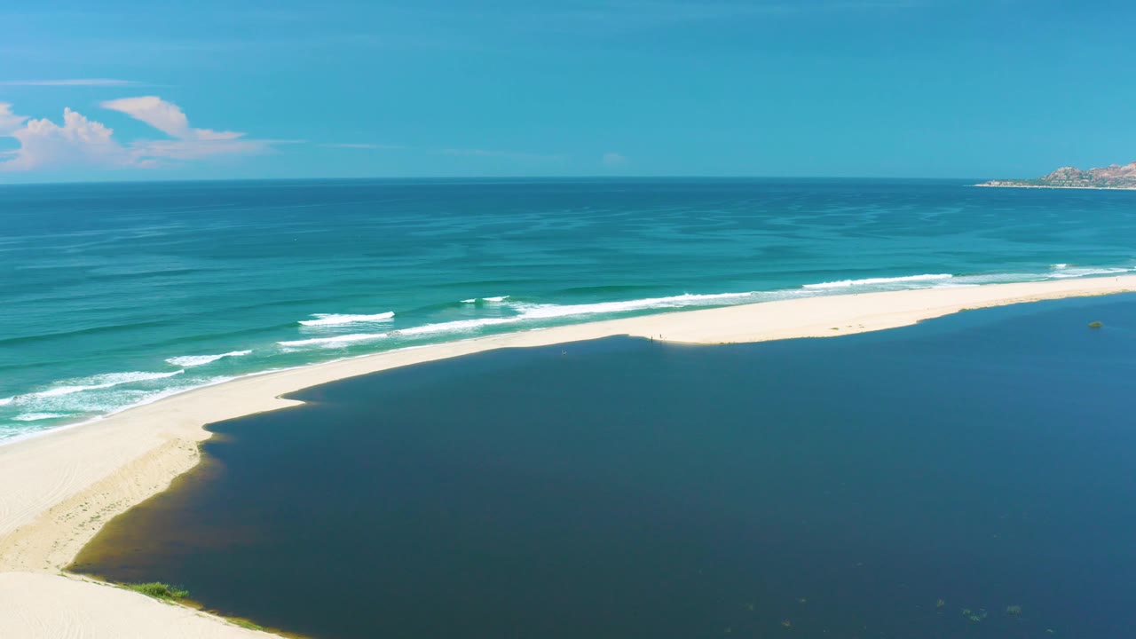 Wide aerial shoreline frame from San Jose Del Cabo showing the sandbar stretching farther right, surf bands near the beach, deep blue foreground water, and no visible people.
