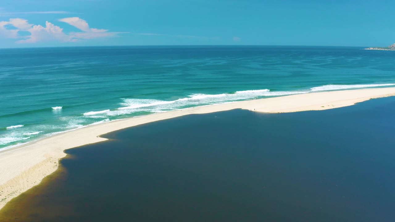 Aerial coastline view in San Jose Del Cabo with surf crossing toward a pale sand spit, darker water below the beach, blue ocean above it, and no visible people.