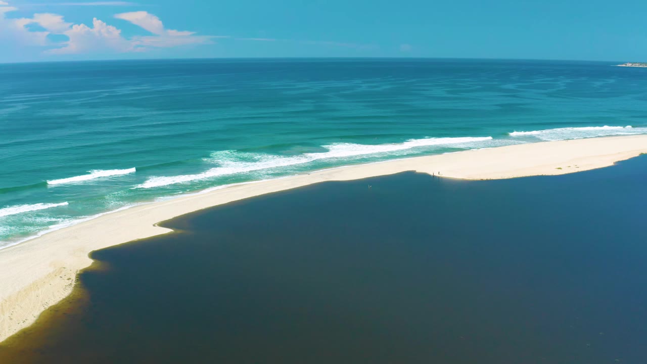 Aerial view in San Jose Del Cabo, Mexico, showing white wave bands curling toward a bright sandbar with dark lagoon water filling the lower frame and no visible people.