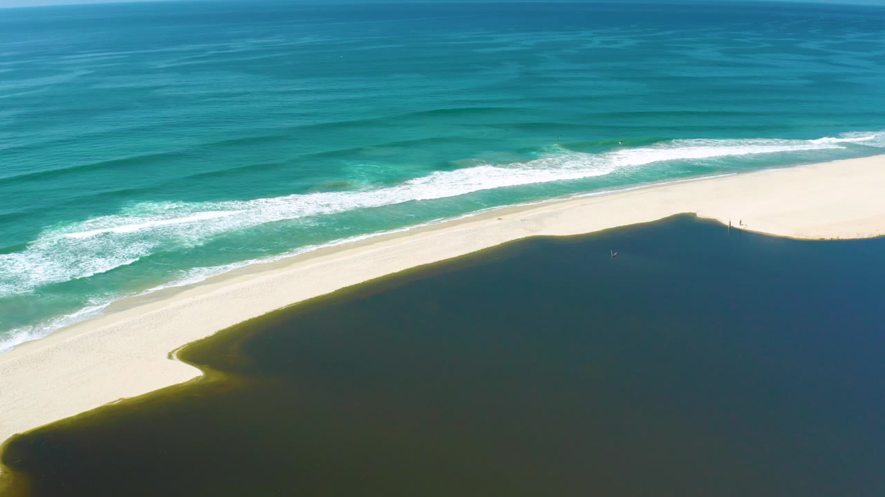 High aerial frame from San Jose Del Cabo, Mexico where foamy surf runs diagonally along the beach next to dark inlet water, with no visible people.