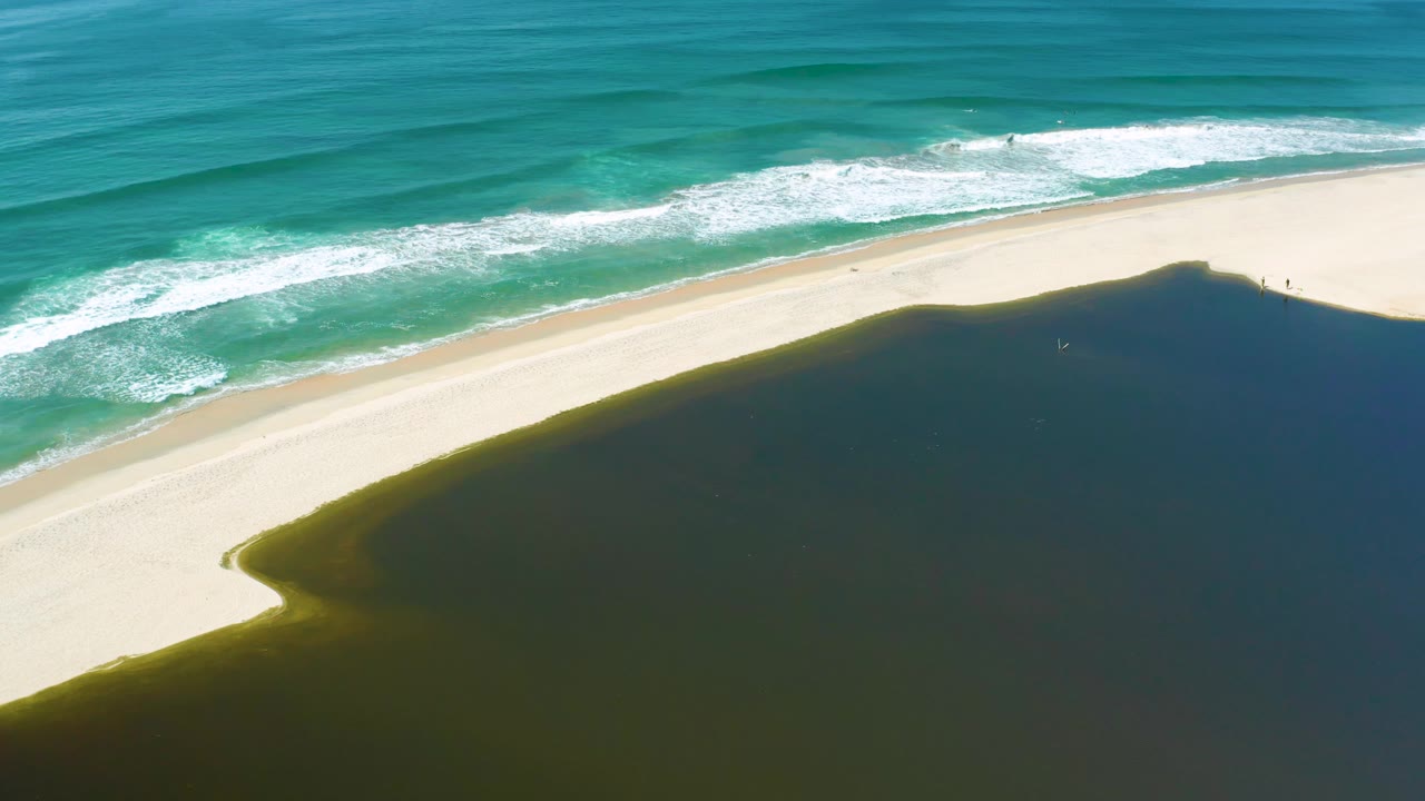Aerial view in San Jose del Cabo, Mexico, with white surf lines near the top, pale sand through the center, and a curved dark water edge below, with no visible people.