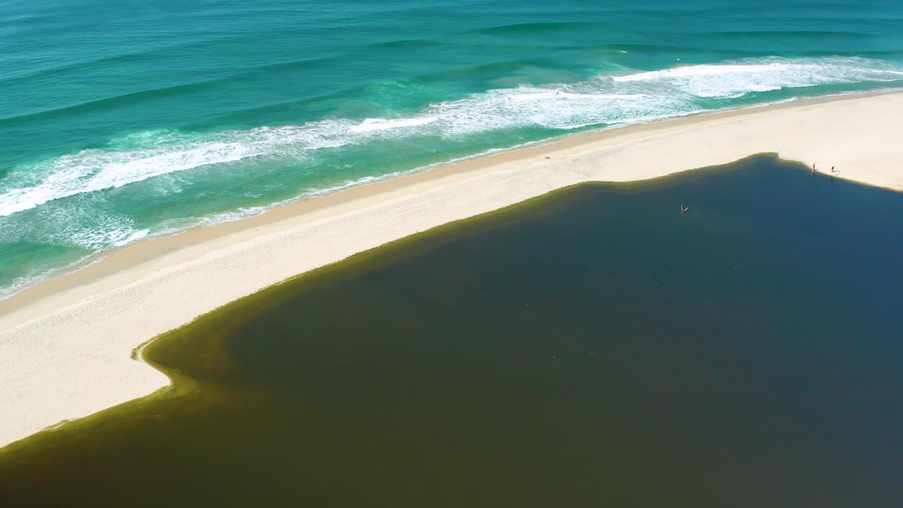 High aerial frame from San Jose del Cabo, Mexico, showing foamy ocean waves, a wide pale beach, and dark green water taking up the lower right, with no visible people.