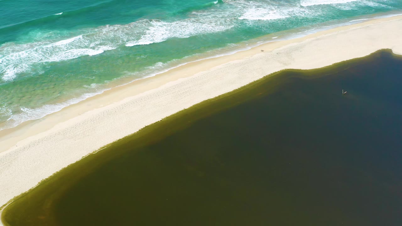 Aerial view in San Jose Del Cabo, Mexico with white wave lines at the top, a bright sand strip across the center, and dark pond water below, with no visible people.