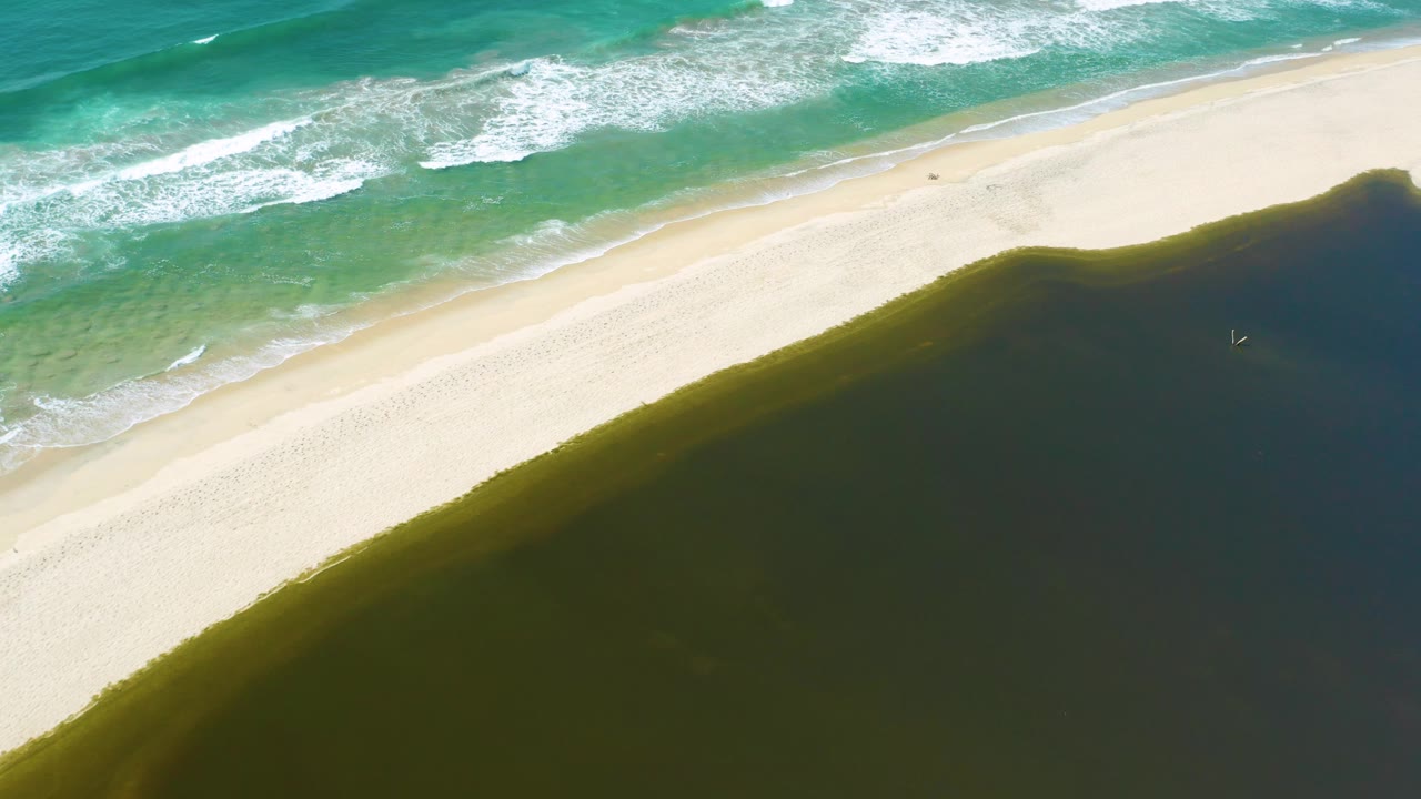 Overhead San Jose Del Cabo coastline frame showing foamy blue waves meeting pale sand while the dark pond fills the lower right, with no visible people.