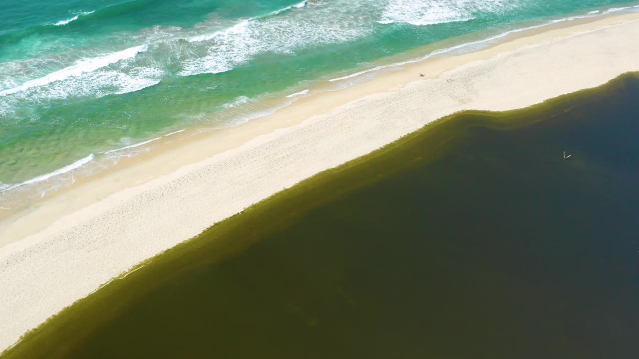Aerial frame in San Jose Del Cabo, Mexico where the bright sandbar curves between white surf at upper left and dark green pond water below, with no visible people.